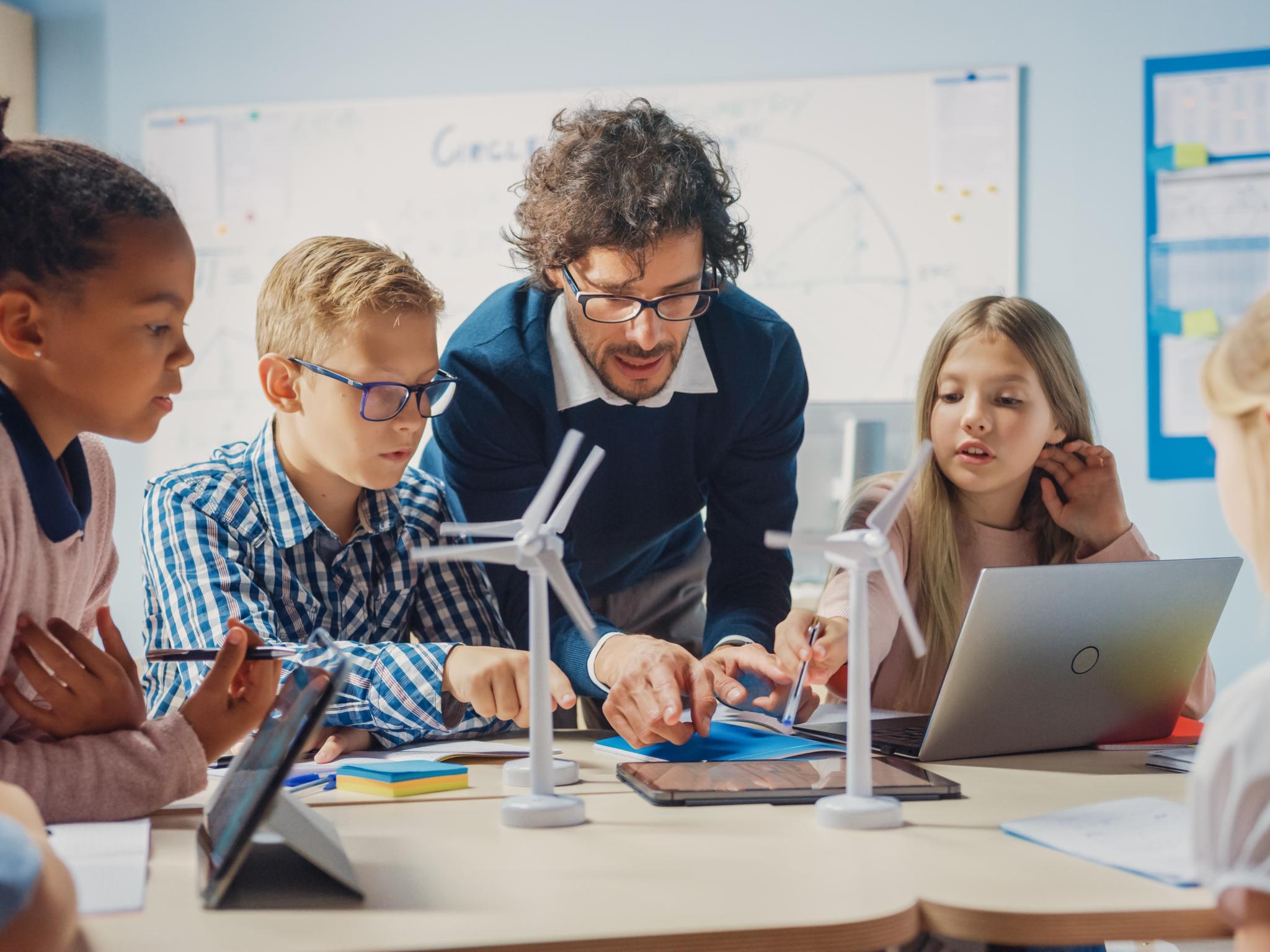 Man sieht Schüler*innen an einem Tisch mit Tablet, Laptop und Miniaturwindrädern. Ein Lehrer beugt sich zur Gruppe, um etwas zu erklären