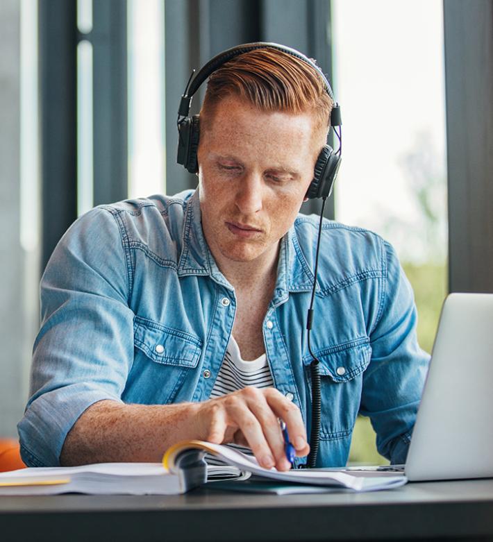 ein Mann welcher Kopfhörer auf hat und an einem Laptop am arbeiten ist ausgeschmückt ist das Bild mit einer lila Sprechblase