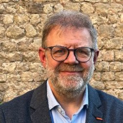 Herr mit Brille steht lächelnd vor einer Natursteinmauer und schaut in die Kamera.