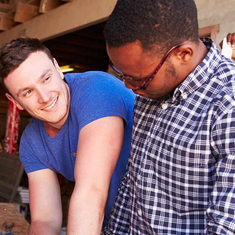 Ein junger Mann erklärt und arbeitet mit 4 farbigen Männern an einer Werkbank. Sie haben Holzherzen gefertigt.