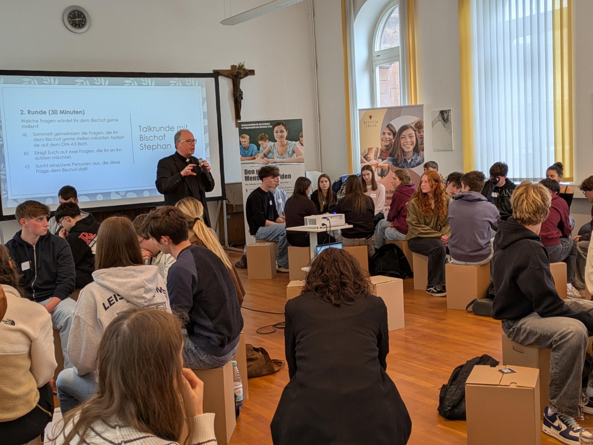 Bischof Ackermann spricht stehend in ein Mikro zwischen vielen Schüler*innen, die auf Hockern in Sitzkreisen versammelt sind.