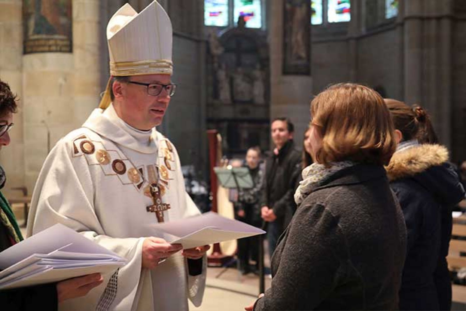 Bischof Dr. Stephan Ackermann steht vor einer jungen Frau, die Teil eines Halbkreises im Dom ist und schaut sie an. in der Hand hält er ein Zertifikat für sie..