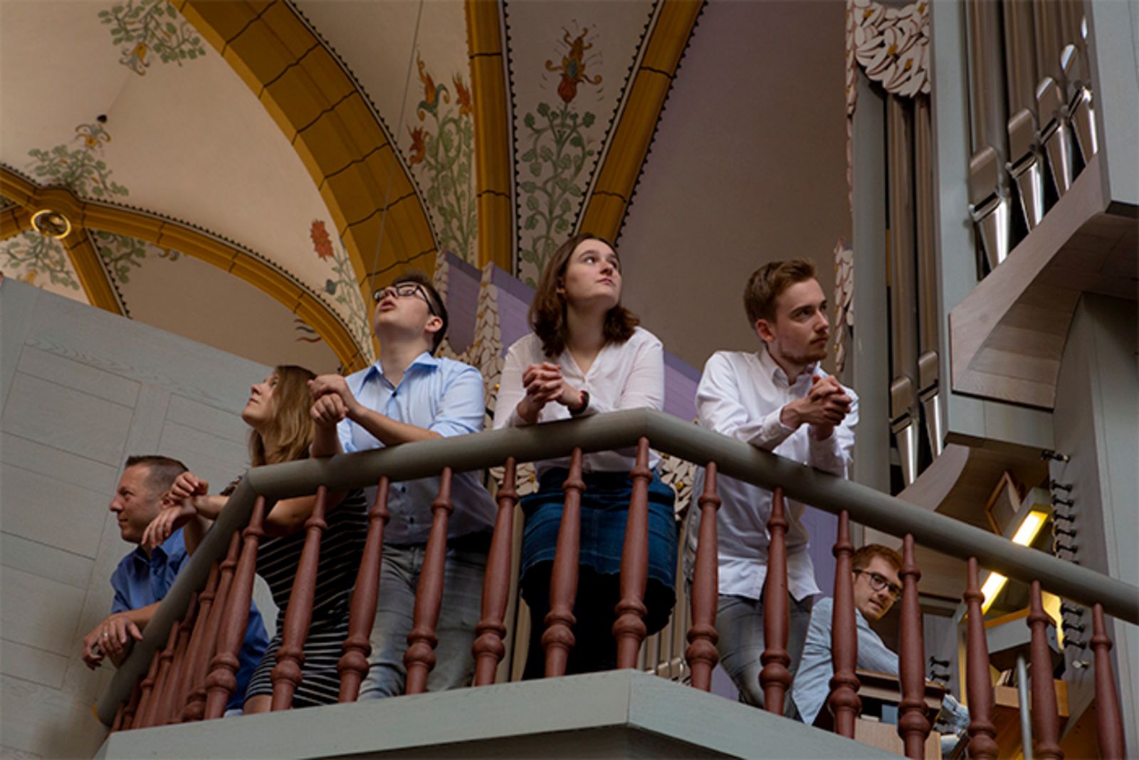 5 Menschen lehnen auf dem Geländer einer Kirchenempore und schauen sich in der Kirche um. Ein Mann spielt dabei Orgel.