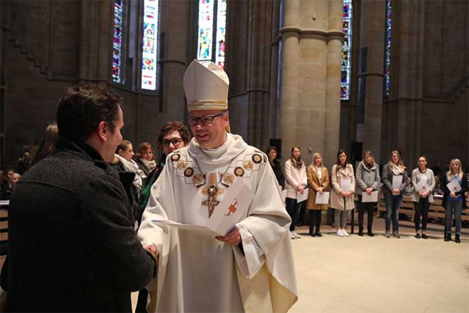 Viele junge Männer und Frauen stehen im Halbkreis im Trier Dom. Bischof Dr. Ackermann reicht einem Mann die Hand und übergibt ihm ein Zertifikat.