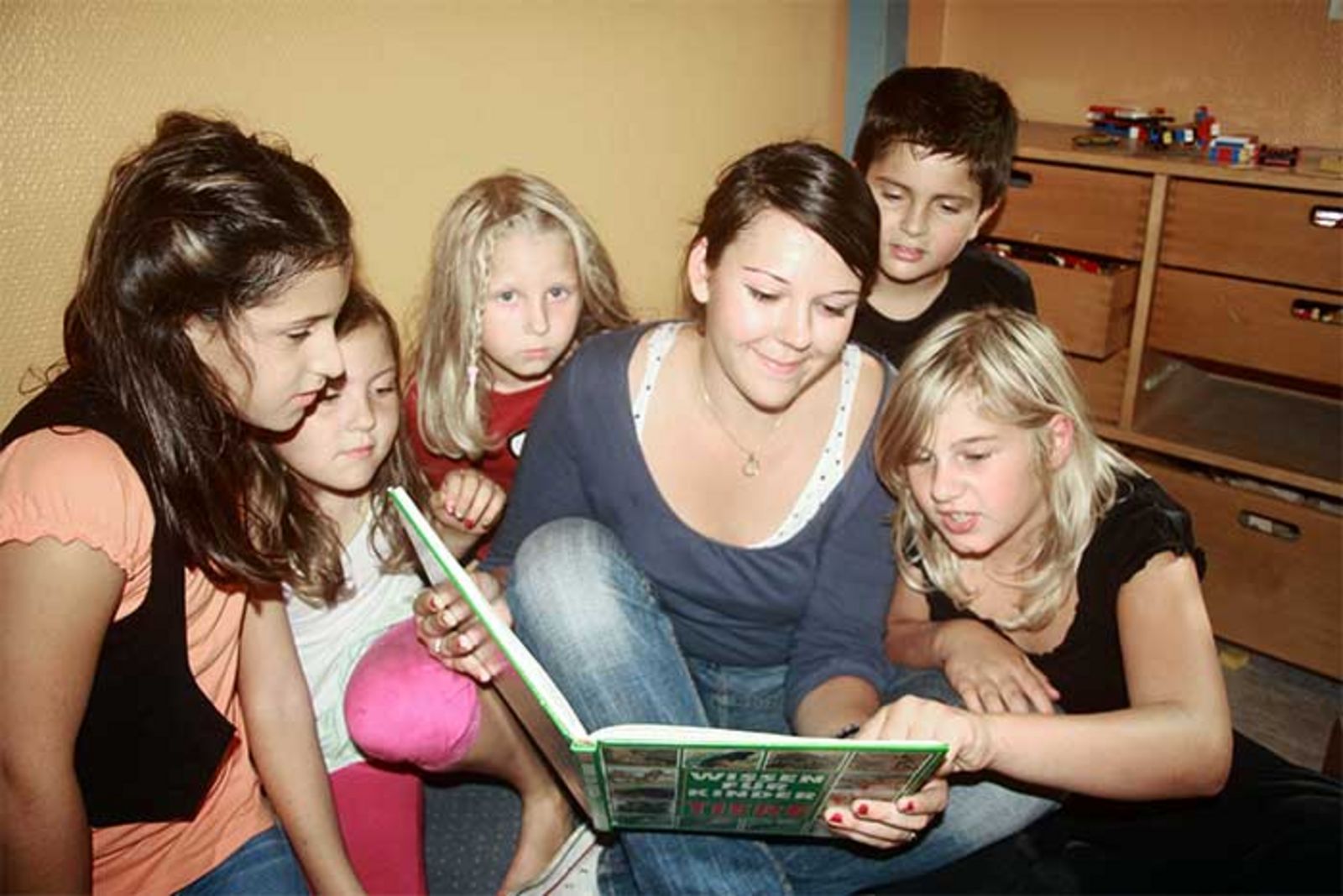 Ein junges Mädchen sitzt mit mehreren Kindern auf dem Fußboden und liest ihnen eine Geschichte vor.