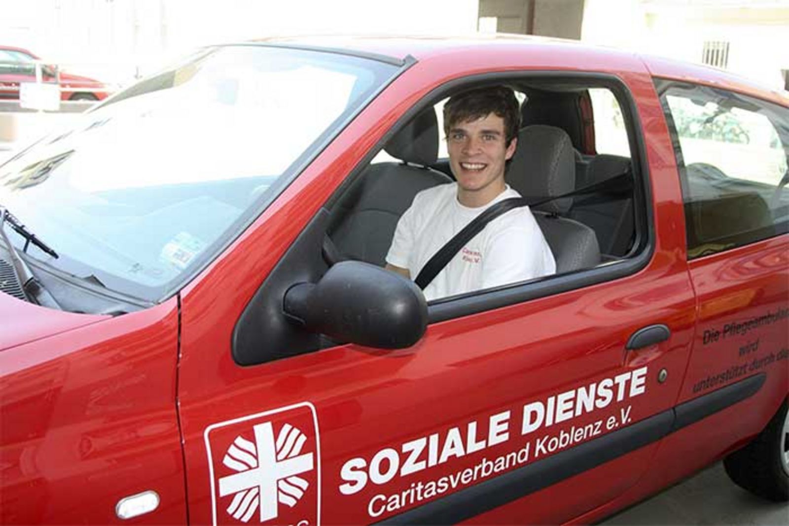 Ein junger Mann sitzt in einem roten Firmenfahrzeug der Sozialen Dienste Caritas Koblenz. 